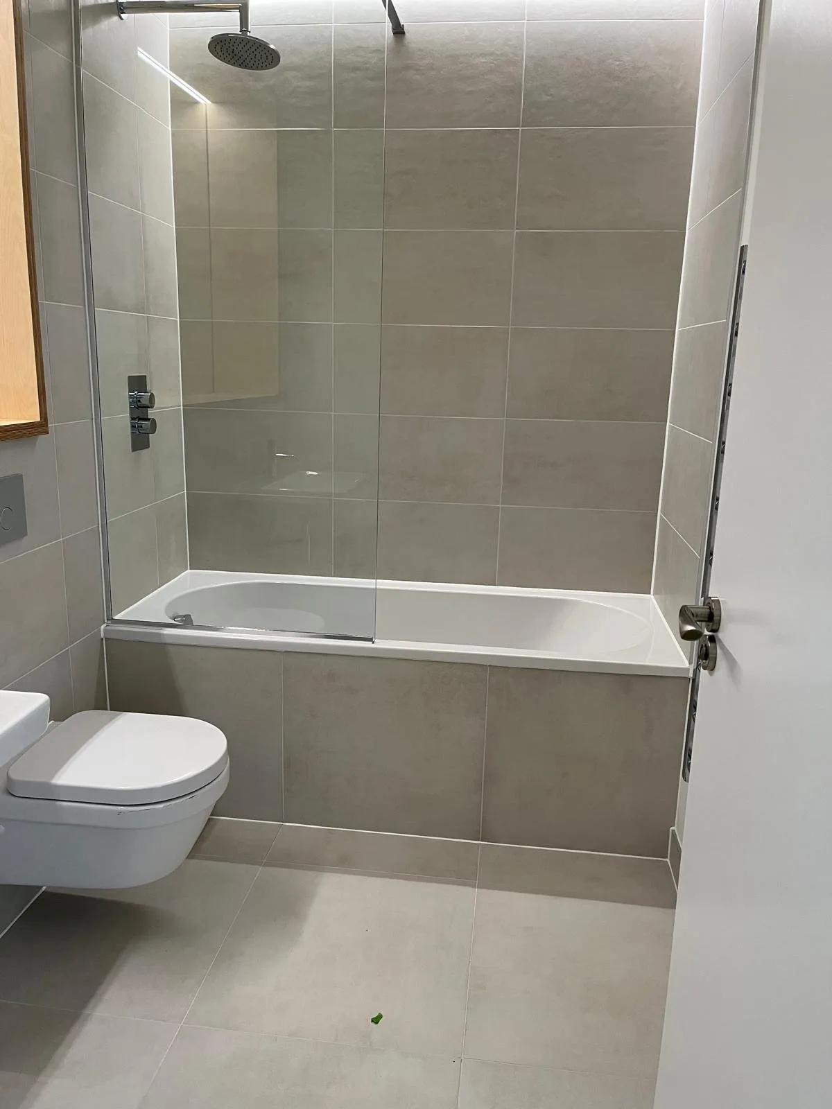 Finished modern bathroom tiled in greige concrete-effect large-format tiles, with a bath shower screen and wall-hung WC