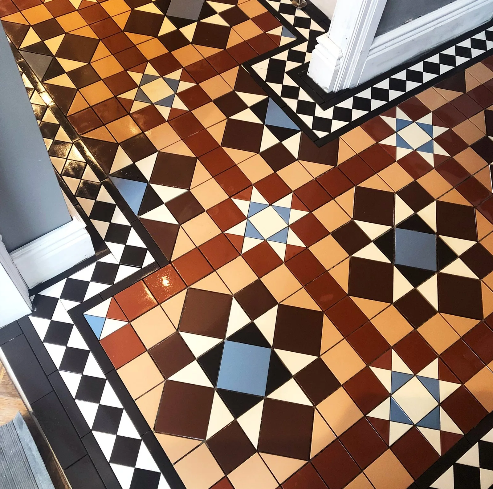 Glossy geometric floor tiling in burnt orange, terracotta, brown, tan and cream with steel-blue centres to black-and-cream star motifs, edged by a black-and-white triangle border