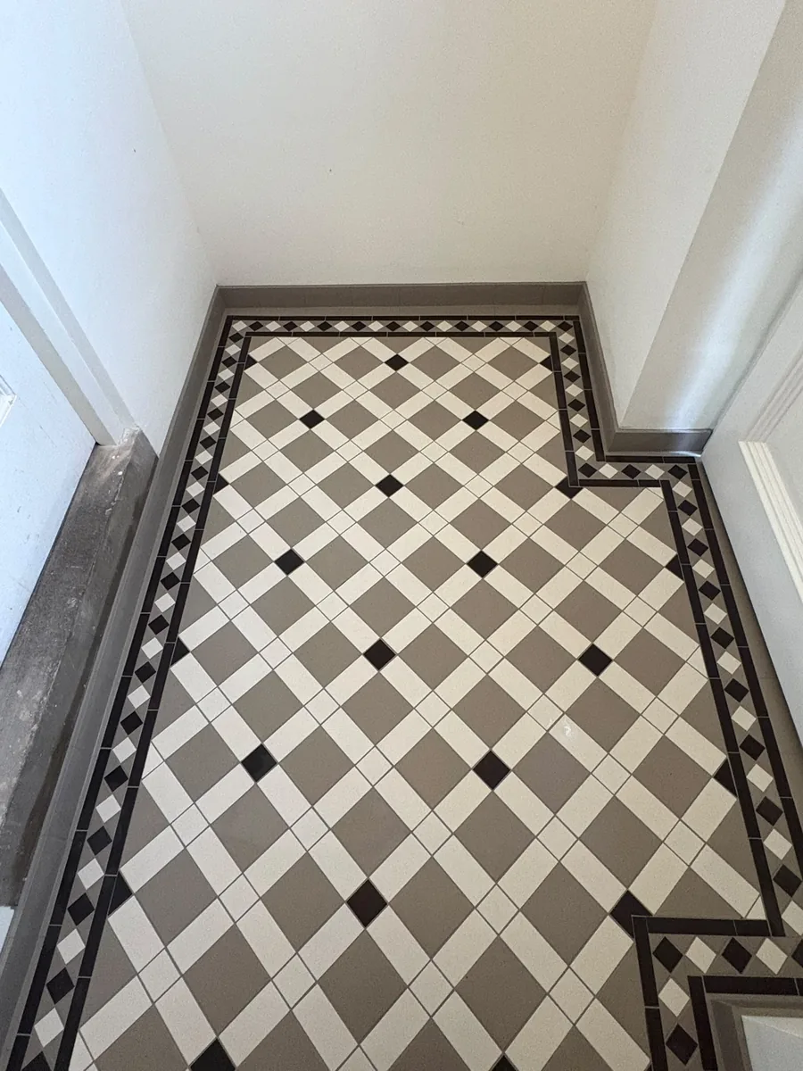Narrow floor in grey and white lattice tiles with small black square insets and a black-and-white diamond border that steps around a wall return, finished with grey tiled skirting