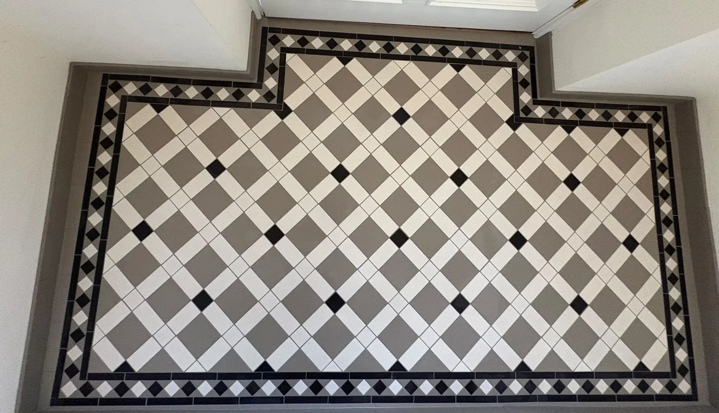 Grey and white tiles laid on the diagonal in a lattice with small black square insets, framed by black bands and a black-and-white diamond border that steps around the shape of the floor
